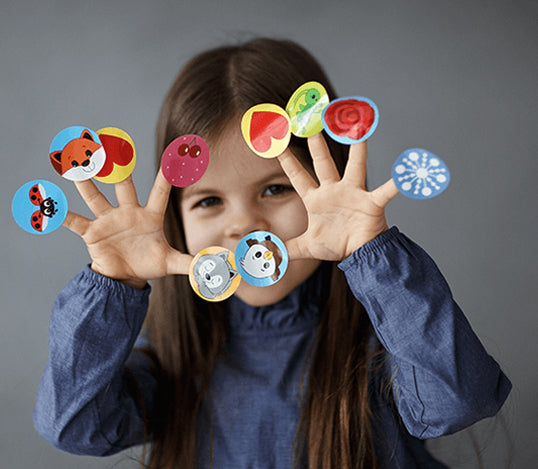 Young girl holding up fingers with colorful animal and fruit stickers on each fingertip against a gray background
