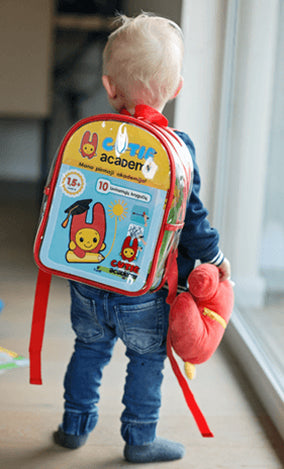 A young child with blonde hair, wearing jeans and a navy sweater, stands indoors facing away, carrying a colorful backpack and holding a red plush toy in one hand.