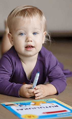 Young child with blue eyes wearing a purple shirt holding a marker and sitting by a colorful book indoors