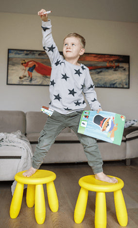 A young boy stands heroically with one foot on each of two yellow stools, holding a marker in one hand raised above his head and an open book in the other, in a living room with a couch and artwork on the wall behind him.